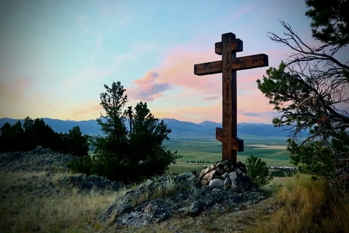 The monastery’s wooden three-bar cross on a rocky rise at dusk, with the valley and mountains beyond.