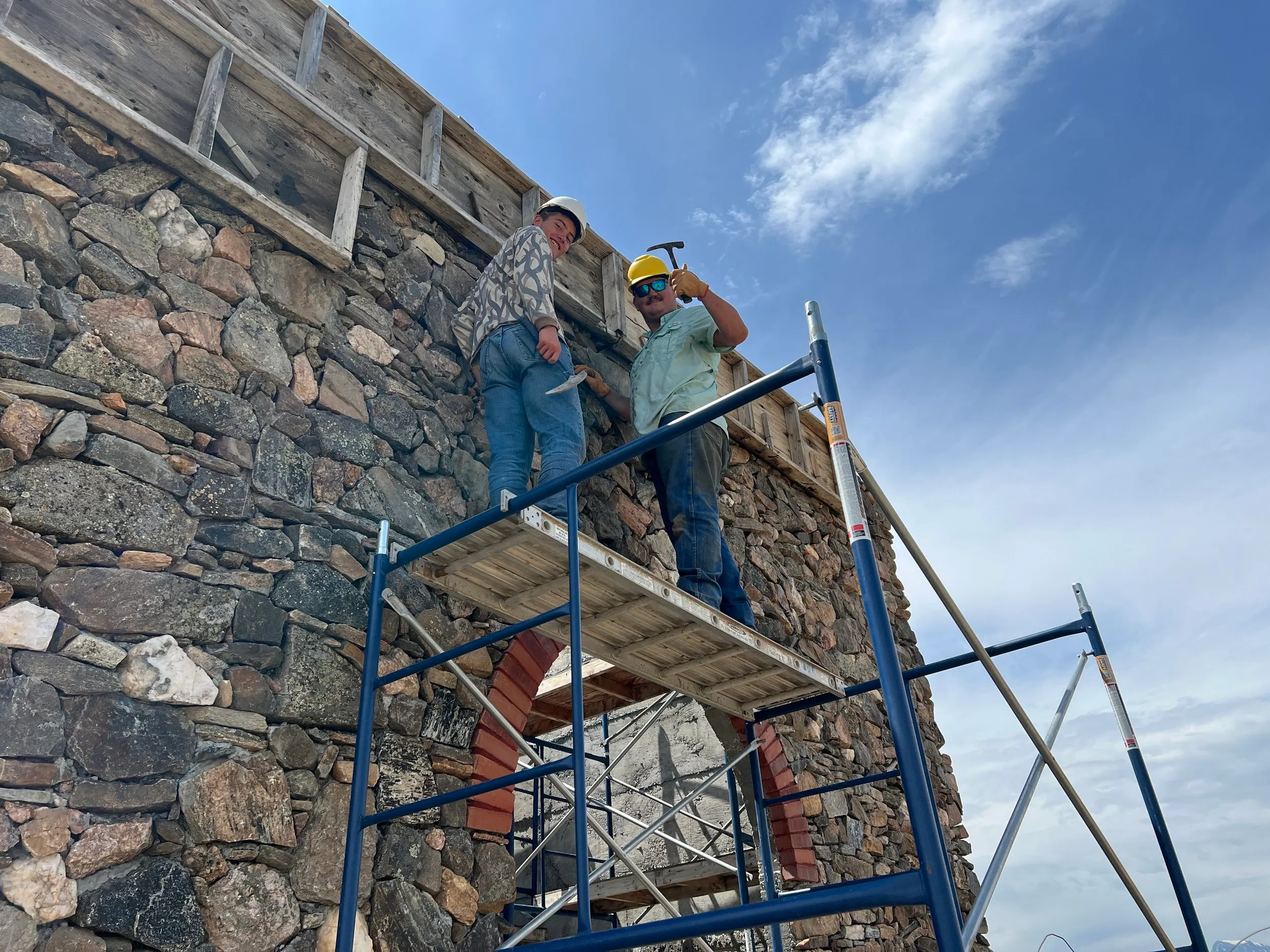 Builders on scaffolding beside the rising stone walls of the monastery’s entrance tower, under a blue sky.
