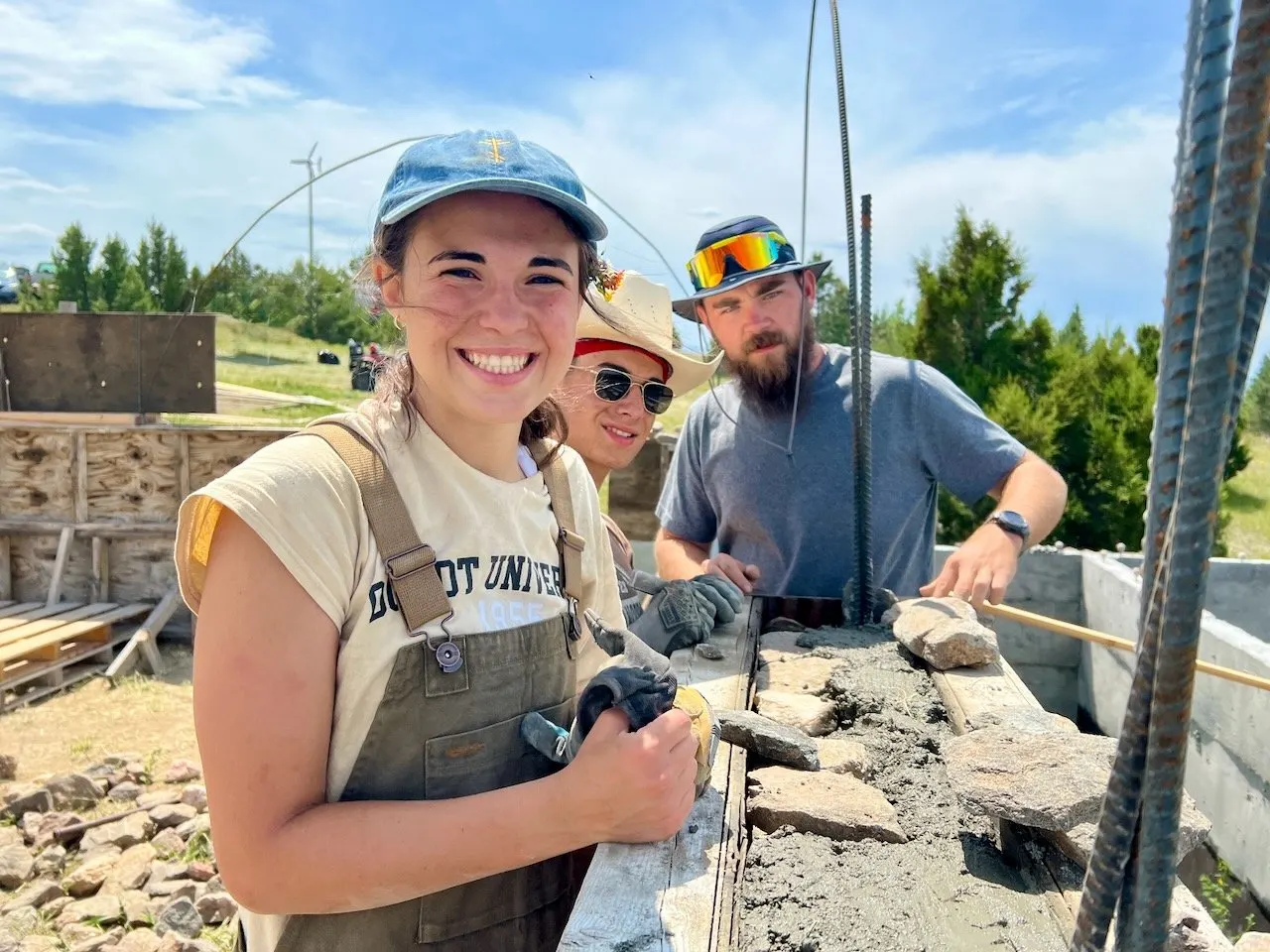 A smiling young woman in a cap and overalls holds a trowel beside a stone wall under construction, with two crew members behind her.