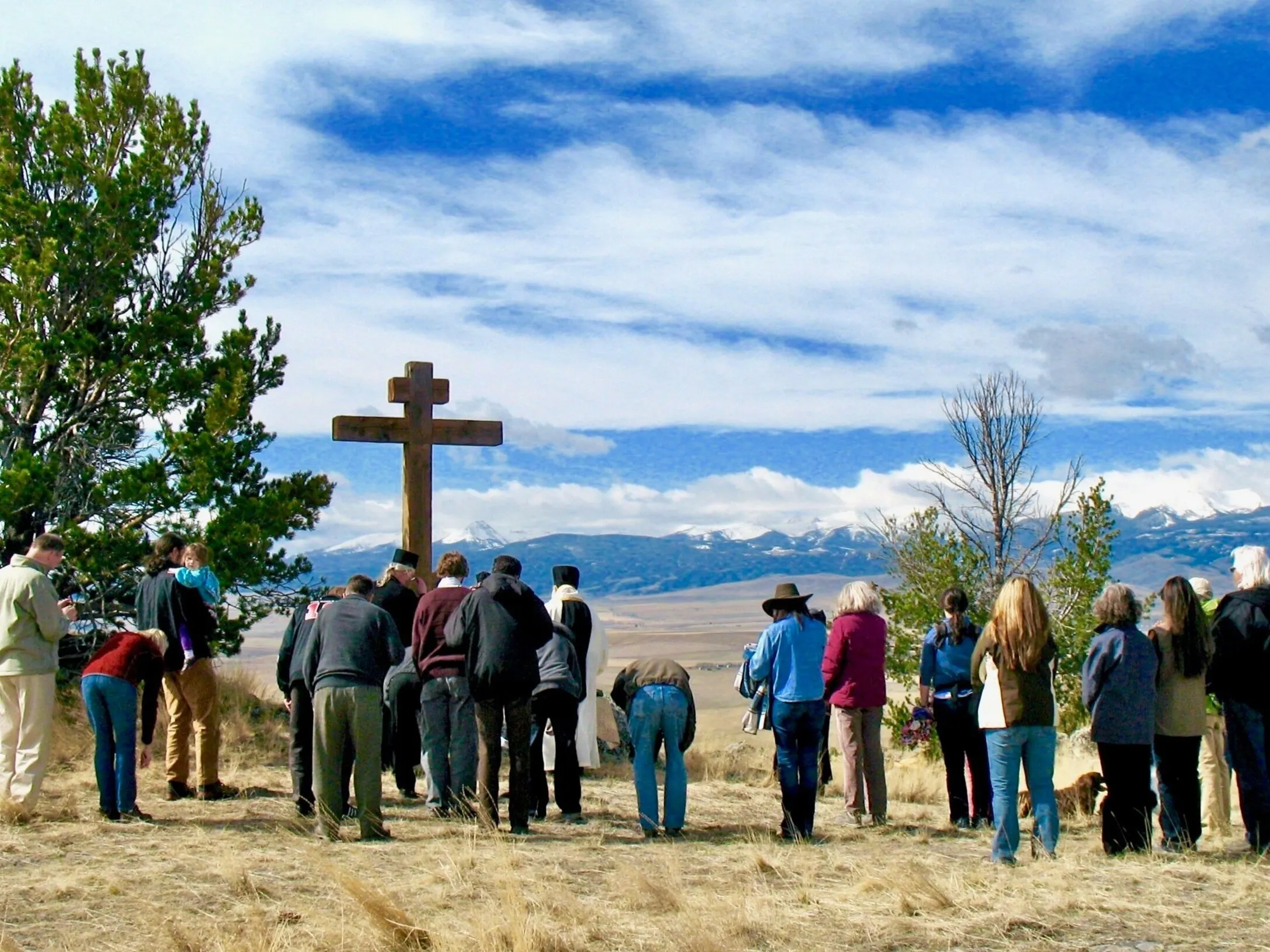 A crowd of the faithful, with clergy in vestments, gathered around a tall wooden cross on a grassy hilltop under a wide sky.