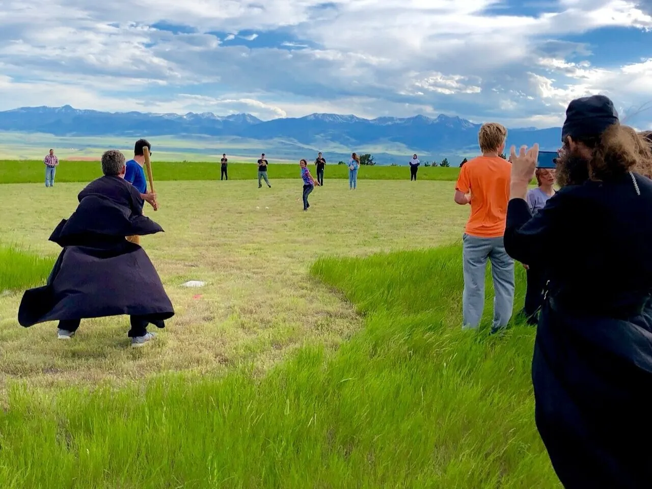 A monk in a black cassock crouches to field a ball during an evening softball game on a grassy field.