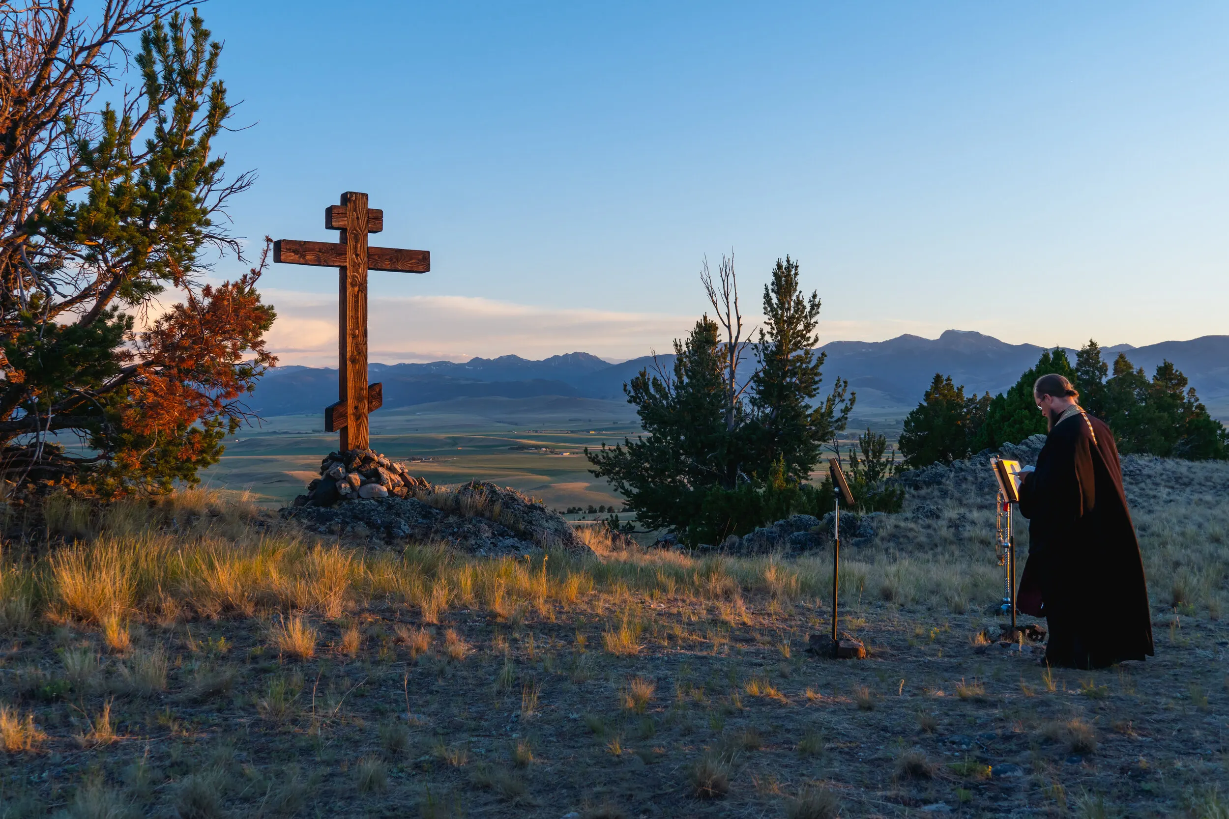 At sunset, a monk reads Vespers at a lectern beside the tall wooden cross overlooking the valley.