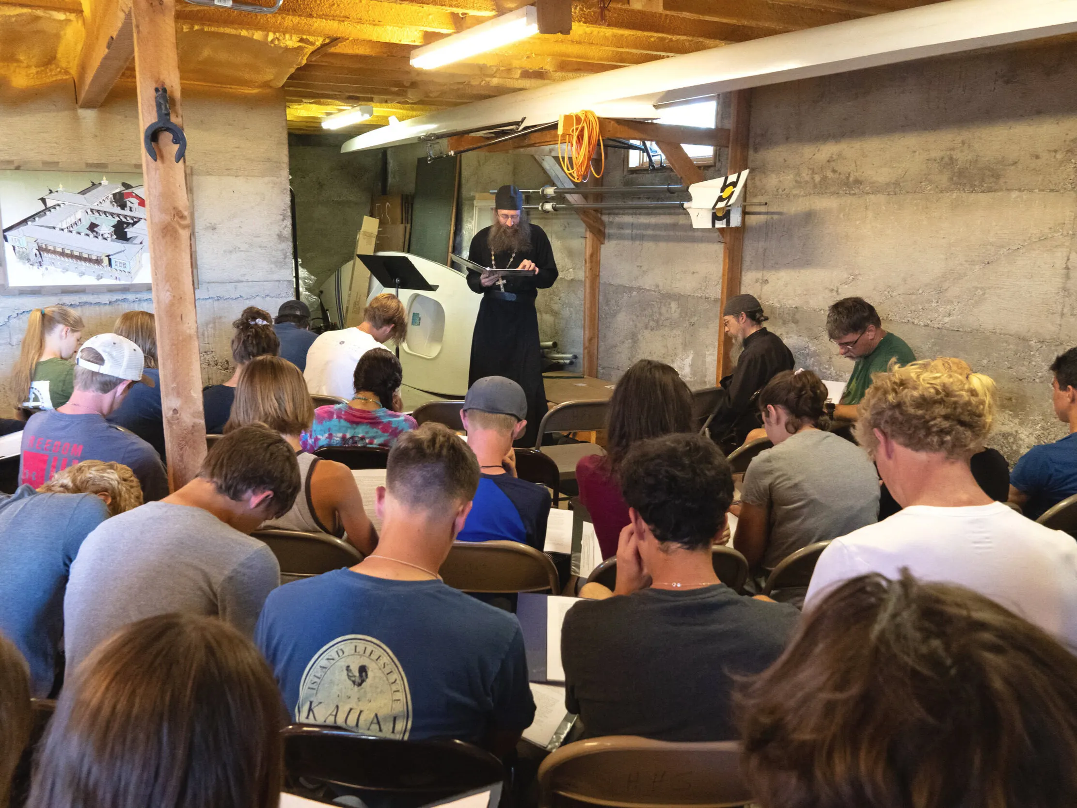 Young Work Week participants seated in rows listen to a monk speaking at the front of a timber-framed hall.