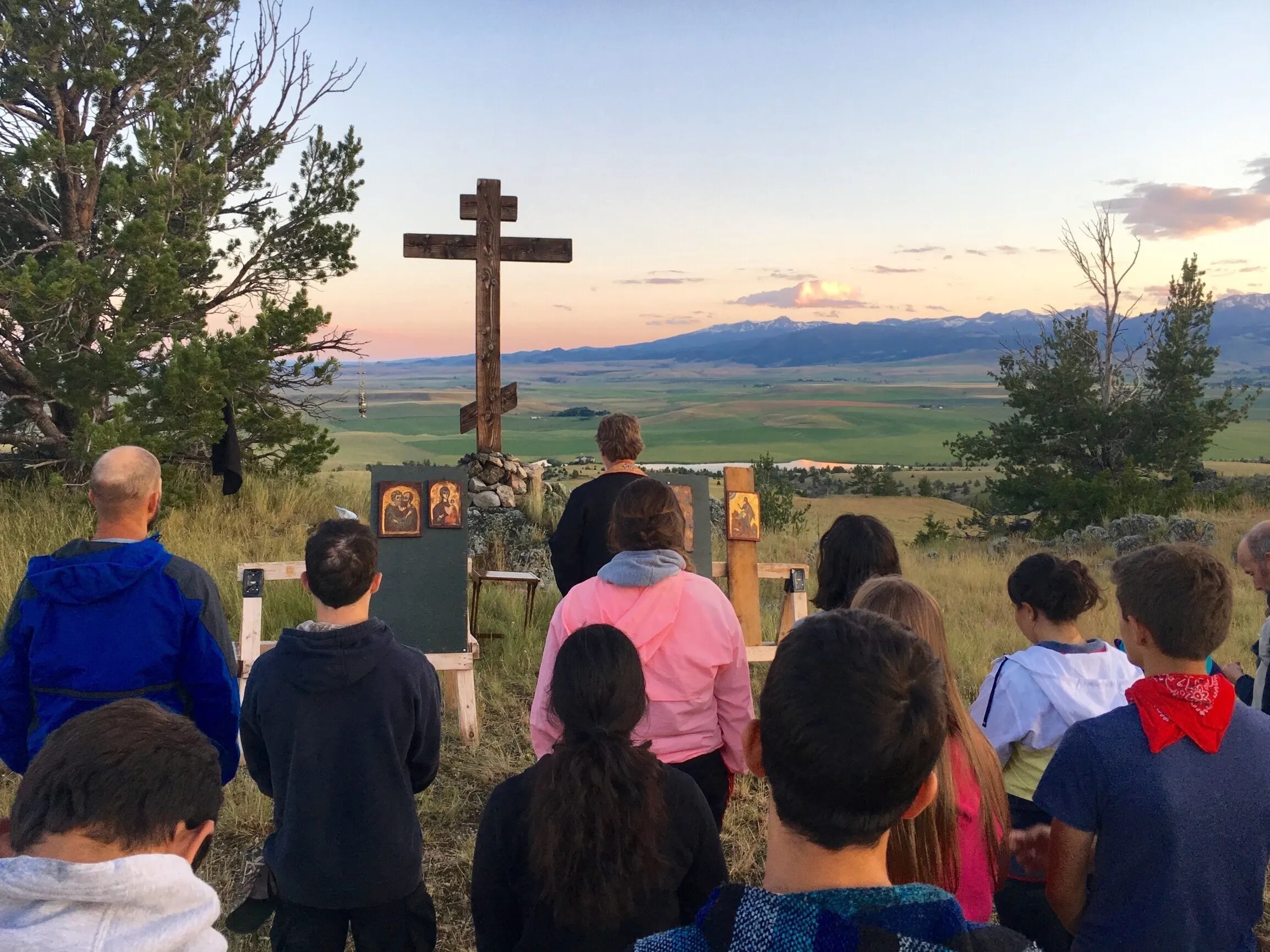 Young people gathered at the tall wooden cross at sunset for Vespers, the valley glowing behind them.